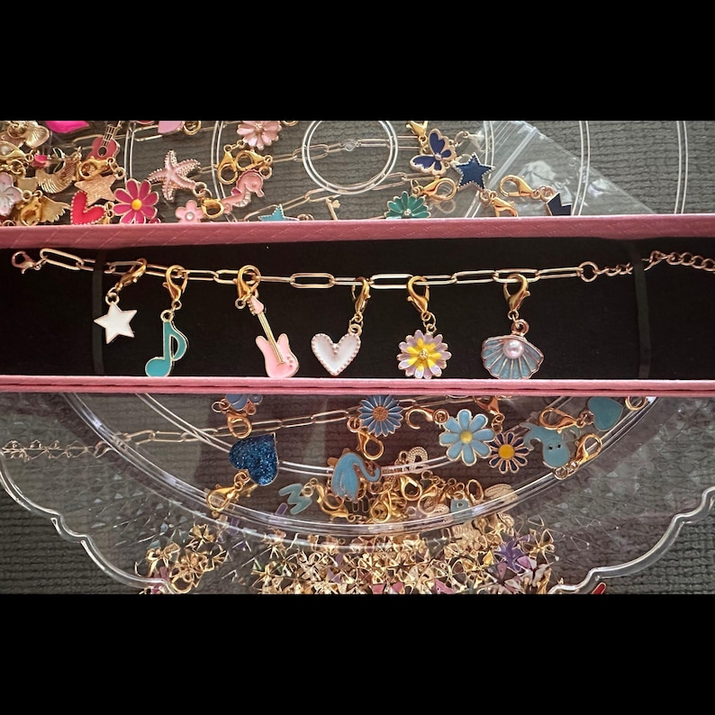 May include: A gold-toned chain bracelet featuring six removable enamel charms: a white star, a teal musical note, a pink electric guitar, a white heart, a multi-colored flower, and a blue seashell with a pearl, displayed in a pink box.
