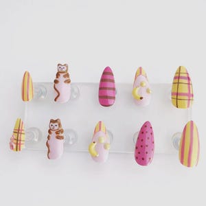 May include: A collection of ten false nails with diverse designs. The nails display patterns including stripes, polka dots, and plaid in pink, yellow, and brown. Some nails have 3D embellishments like a cartoon bear and bananas.