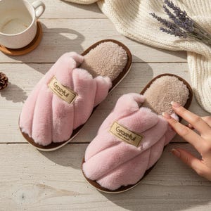 Può includere: Un paio di pantofole rosa in peluche con suola marrone e interno beige. Le pantofole hanno un'etichetta con la scritta "Graceful". Le pantofole sono su una superficie di legno chiaro.