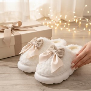 May include: A pair of fluffy white slippers with a cream-colored bow and a thick white sole. The slippers are next to a gift box with a cream-colored ribbon. The background includes fairy lights and a window.