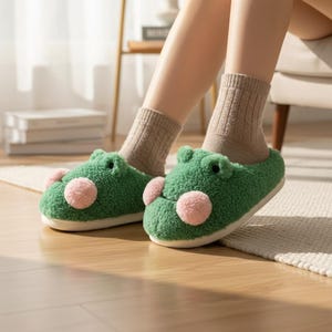 Può includere: Pantofole verdi a forma di rana con accenti rosa sulle guance. Le pantofole hanno una consistenza soffice e sono indossate con calzini color beige. Le pantofole sono su un pavimento in legno chiaro.