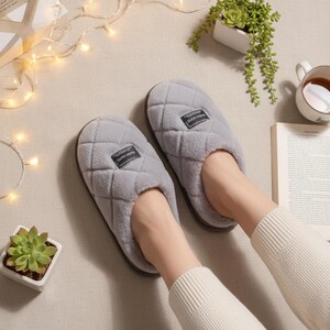 May include: A pair of gray quilted slippers with a soft, plush texture. Each slipper has a small rectangular label. The slippers are designed for indoor use and provide comfort and warmth. The slippers are shown on a neutral surface.