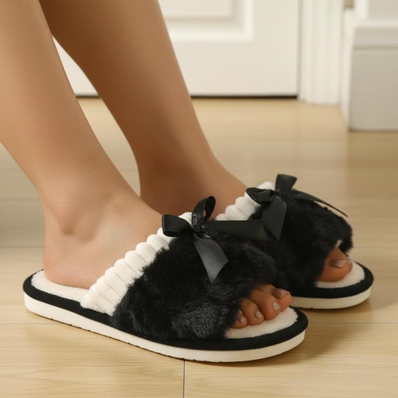 Pu&ograve; includere: Pantofole nere e bianche a punta aperta con un design soffice e un fiocco nero. Le pantofole hanno un bordo bianco a coste e una suola nera. L'immagine mostra le pantofole indossate.