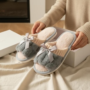 Pu&ograve; includere: Un paio di pantofole grigie e rosa a punta aperta con un dettaglio a fiocco. Le pantofole hanno una parte superiore grigia soffice, un interno rosa e una fascia a maglia beige chiaro. Le pantofole sono presentate in una scatola bianca.