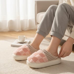 Può includere: Pantofole rosa e bianche a quadri con una spessa suola bianca. Le pantofole hanno una consistenza morbida e soffice e un design a coste sulla parte superiore. L'immagine mostra le pantofole indossate in casa.
