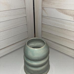 Może przedstawiać: Ręcznie wykonany, jasnoniebieski ceramiczny wazon o unikalnym, żebrowanym wzorze. Wazon ma zaokrągloną górę i szereg poziomych żeber, które tworzą teksturowany wygląd. Jest prezentowany na białej powierzchni na tle drewnianych okiennic.