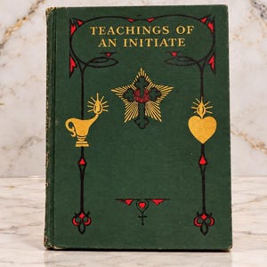 Op de afbeelding: Een donkergroen hardcover boek met de titel Teachings of an Initiate. De kaft toont een gouden ster met een kruis, een gouden olielamp en een gouden hart. Decoratieve zwarte en rode lijntekeningen omlijsten de illustraties op de voorzijde.