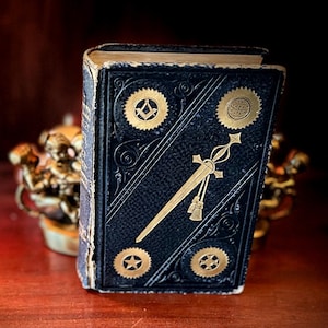 Puede incluir: Un libro de tapa dura negro con detalles dorados en relieve en la portada, que incluyen un emblema central de espada, un símbolo de escuadra y compás, un globo terráqueo, una estrella y una cruz. El lomo muestra bordes desgastados.