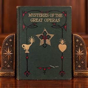 May include: A dark green hardcover book titled Mysteries of the Great Operas with gold and red decorative illustrations on the front cover, positioned between two dark bronze-colored metal bookends featuring star and celestial motifs.