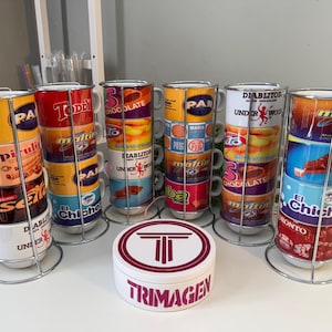May include: Stacked coffee mugs with colorful designs and text, held in metal racks. The mugs feature various brand logos and graphics. A white container with the word "TRIMAGEN" is in the foreground.