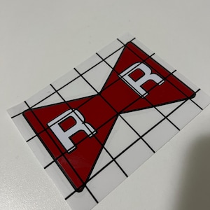 May include: A red and white sticker featuring a stylized bow tie design with the letter "R" in white on each side. The sticker is on a white background with a black grid pattern.