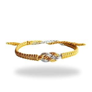 Puede incluir: Una pulsera de la amistad dorada y plateada con un diseño de nudo. La pulsera está hecha de cordón tejido y presenta un cierre de nudo corredizo para un ajuste ajustable. El nudo es una mezcla de hilos dorados y plateados.