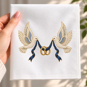 Op de afbeelding: Een vierkant wit stoffen servet met een geborduurd ontwerp van twee crèmekleurige duiven met blauw getinte vleugels. De duiven houden een donkerblauw lint in hun snavel dat door twee in elkaar grijpende goudkleurige ringen is gestoken.