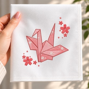May include: A square white fabric handkerchief featuring an embroidered pink origami crane at the center. The crane is decorated with small white floral accents, and the design is surrounded by several embroidered pink cherry blossom flowers.
