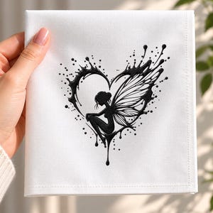May include: A square white fabric napkin featuring a black embroidered design of a seated fairy with wings positioned inside a heart shape that appears to be formed by liquid ink splatters and drips, creating a high-contrast graphic motif.