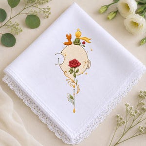 Può includere: Un fazzoletto quadrato bianco con bordo in pizzo caratterizzato da un ricamo raffigurante una piccola figura dai capelli gialli, una volpe e una rosa rossa. Il ricamo include linee decorative dorate, piccole stelle e un gambo verde.