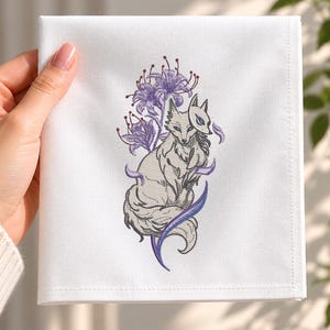 May include: A white square fabric napkin featuring an embroidered design of a grey fox wearing a white mask, surrounded by purple spider lily flowers and swirling purple accents. The embroidery is centered on the smooth white textile surface.