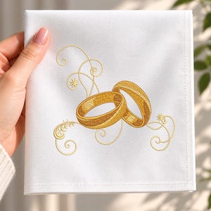 Puede incluir: Una servilleta de tela blanca cuadrada con un diseño bordado de dos anillos dorados entrelazados. Los anillos están rodeados por delicados patrones de filigrana dorada en espiral que se extienden hacia afuera sobre la tela blanca lisa.