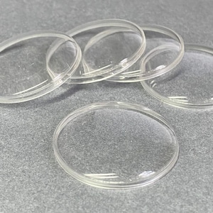 May include: A collection of five clear, circular, domed glass cabochons with smooth, polished edges. Each piece features a transparent, convex surface that creates a magnifying effect, arranged in a cluster to show their uniform shape and clarity.