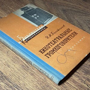 May include: Vintage book with a tan cover and blue spine, featuring Russian text and a black and white illustration. The title is in bold, dark letters. The book is lying on a wooden surface.
