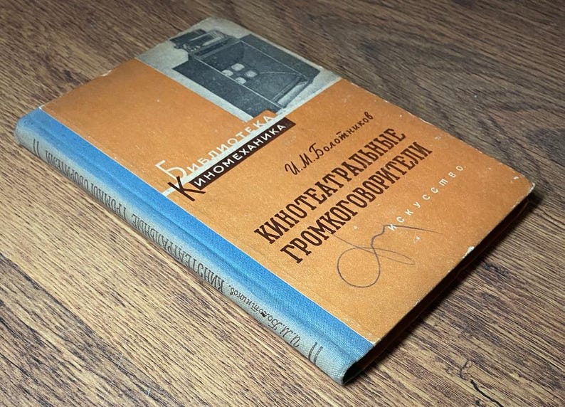 May include: Vintage book with a tan cover and blue spine, featuring Russian text and a black and white illustration. The title is in bold, dark letters. The book is lying on a wooden surface.