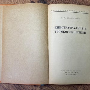 May include: An open vintage book with aged, tan-colored pages. The right page features black Cyrillic text, including the title "Kino-teatral'nye Gromkogovoriteli." The book's spine is orange, and the cover is not visible. The book is on a wooden surface.