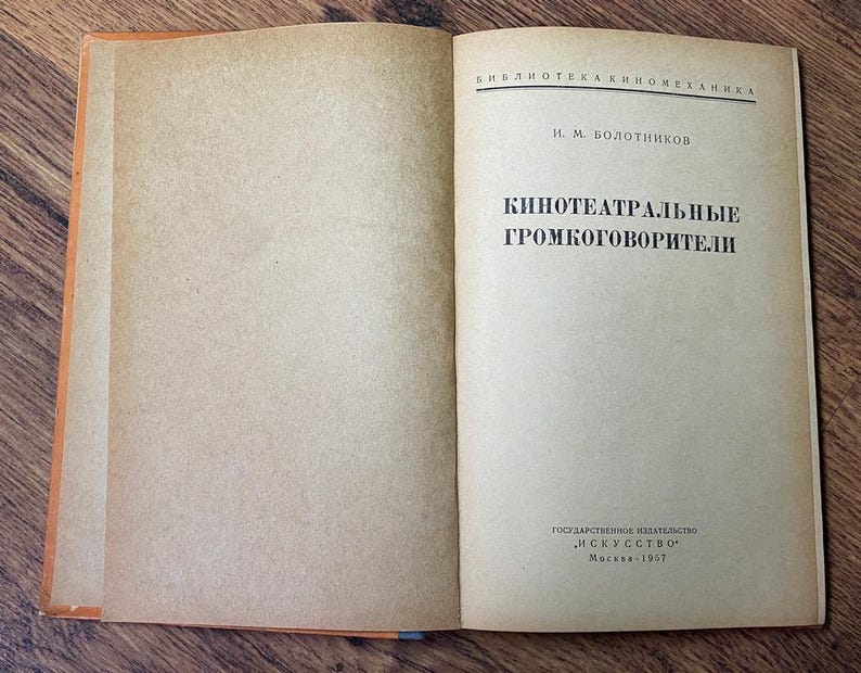 May include: An open vintage book with aged, tan-colored pages. The right page features black Cyrillic text, including the title "Kino-teatral'nye Gromkogovoriteli." The book's spine is orange, and the cover is not visible. The book is on a wooden surface.