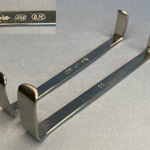 May include: Two silver surgical retractors. Each retractor has a long, flat handle with a curved, rectangular end. The handles are marked with numbers and symbols. The top left corner shows a close-up of the markings, including the initials "B.M."