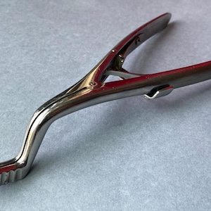 K&ouml;nnte beinhalten: Gl&auml;nzendes, silberfarbenes chirurgisches Instrument mit gebogenem Griff und gezahntem Kiefer. Das Werkzeug besteht aus Metall und scheint in gutem Zustand zu sein. Das Instrument ist f&uuml;r den medizinischen Gebrauch bestimmt.