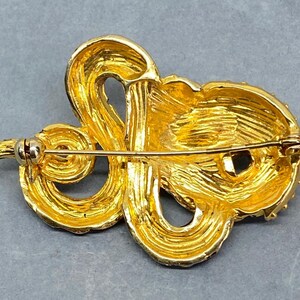 May include: A gold-toned brooch with an ornate, swirling design. The brooch features a textured surface and a pin closure on the back. The design is reminiscent of a stylized flower or abstract shape, with a bright, reflective finish.