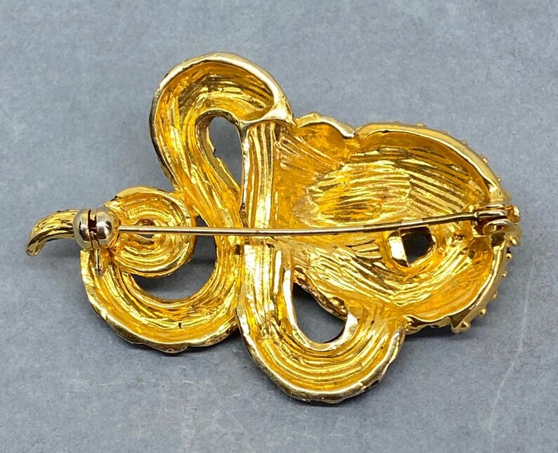 May include: A gold-toned brooch with an ornate, swirling design. The brooch features a textured surface and a pin closure on the back. The design is reminiscent of a stylized flower or abstract shape, with a bright, reflective finish.