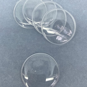 May include: A collection of five clear, circular, dome-shaped glass cabochons. These transparent, smooth, and rounded pieces are arranged with four overlapping in a cluster at the top and one single piece positioned below them.