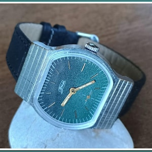 Könnte beinhalten: Eine silberne Armbanduhr mit grünem Zifferblatt und schwarzem Lederarmband. Das Zifferblatt hat goldene Zeiger und ein goldenes "Raketa"-Logo.