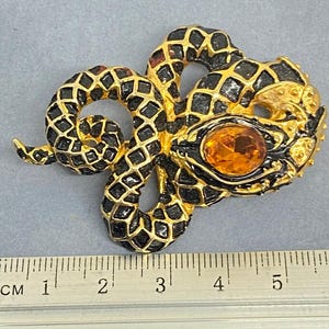 May include: A gold-toned snake brooch with black enamel detailing and an amber-colored gemstone eye. The snake is coiled, with a textured pattern on its body. The brooch measures approximately 2 inches in length.