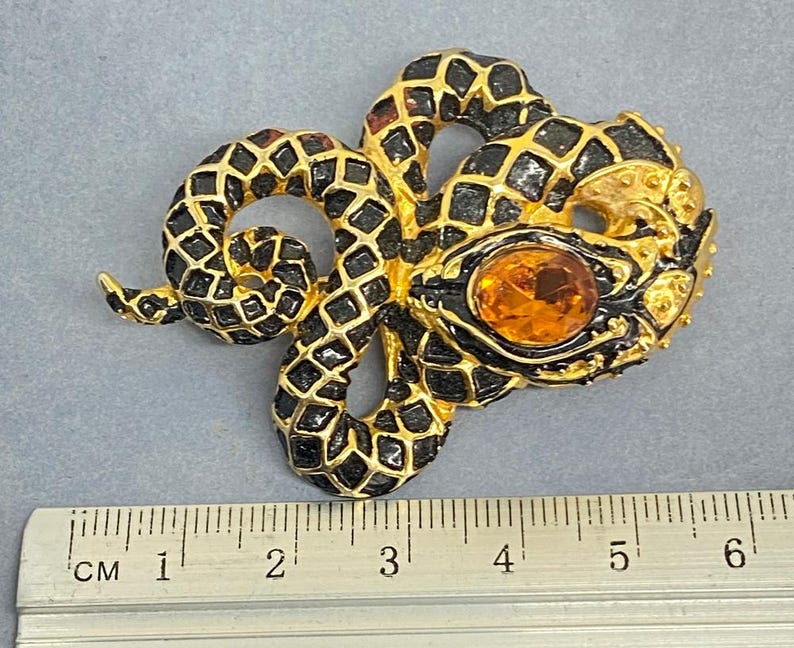 May include: A gold-toned snake brooch with black enamel detailing and an amber-colored gemstone eye. The snake is coiled, with a textured pattern on its body. The brooch measures approximately 2 inches in length.