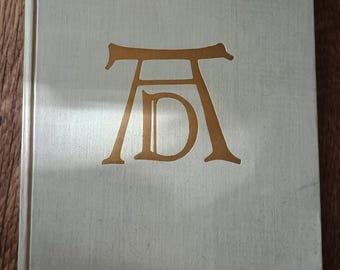 Puede incluir: Un libro gris claro con un diseño en relieve dorado en la portada. El diseño es una letra estilizada "A" con una "D" dentro de la "A".