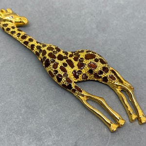 K&ouml;nnte beinhalten: Goldfarbene Giraffenfigur mit braunen Flecken. Die Giraffe hat einen langen Hals und Beine sowie einen kleinen Kristall auf dem Kopf. Die Figur liegt auf einer grauen Oberfl&auml;che.
