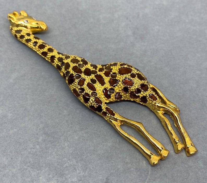 K&ouml;nnte beinhalten: Goldfarbene Giraffenfigur mit braunen Flecken. Die Giraffe hat einen langen Hals und Beine sowie einen kleinen Kristall auf dem Kopf. Die Figur liegt auf einer grauen Oberfl&auml;che.