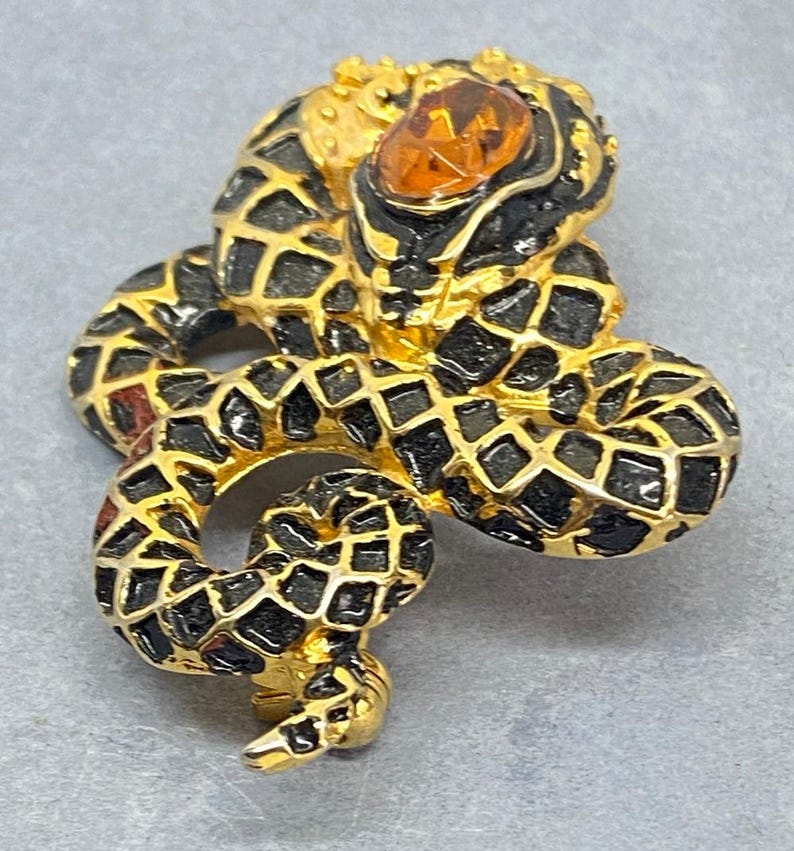 May include: A gold-toned snake brooch with black enamel detailing and a large amber-colored stone eye. The snake's body is coiled, and the design features a scale pattern.