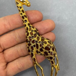K&ouml;nnte beinhalten: Goldfarbene Giraffenbrosche mit braunen und bernsteinfarbenen Akzenten. Die Giraffe hat einen langen Hals und Beine mit einem strukturierten Muster auf ihrem K&ouml;rper. Die Brosche hat einen Nadelverschluss auf der R&uuml;ckseite.