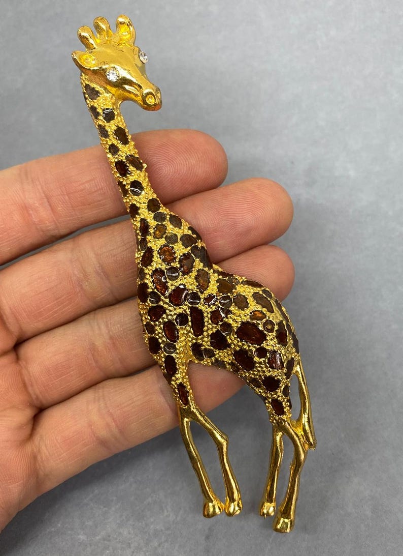 K&ouml;nnte beinhalten: Goldfarbene Giraffenbrosche mit braunen und bernsteinfarbenen Akzenten. Die Giraffe hat einen langen Hals und Beine mit einem strukturierten Muster auf ihrem K&ouml;rper. Die Brosche hat einen Nadelverschluss auf der R&uuml;ckseite.