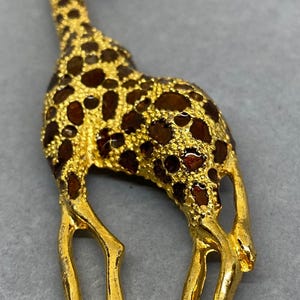 K&ouml;nnte beinhalten: Goldfarbene Giraffenbrosche mit braunen Emaille-Flecken. Die Giraffe steht, mit detailliertem langem Hals und Beinen. Die Brosche ist wahrscheinlich aus Metall und ein dekoratives Accessoire.