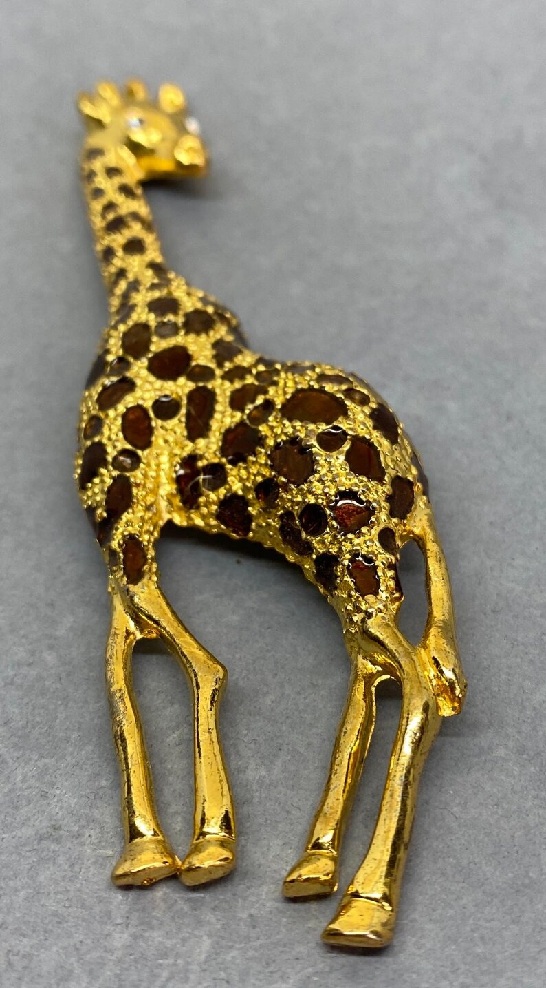 K&ouml;nnte beinhalten: Goldfarbene Giraffenbrosche mit braunen Emaille-Flecken. Die Giraffe steht, mit detailliertem langem Hals und Beinen. Die Brosche ist wahrscheinlich aus Metall und ein dekoratives Accessoire.