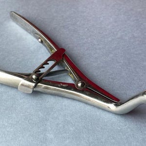 K&ouml;nnte beinhalten: Ein silberfarbenes chirurgisches Instrument mit rotem Akzent in der Mitte. Das Werkzeug hat gebogene, spitze Enden und einen Griff mit geriffelter Oberfl&auml;che. Das Instrument ist f&uuml;r den medizinischen Gebrauch bestimmt.