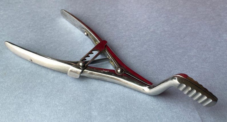 K&ouml;nnte beinhalten: Ein silberfarbenes chirurgisches Instrument mit rotem Akzent in der Mitte. Das Werkzeug hat gebogene, spitze Enden und einen Griff mit geriffelter Oberfl&auml;che. Das Instrument ist f&uuml;r den medizinischen Gebrauch bestimmt.