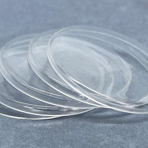 Puede incluir: Una pila de discos de plástico transparentes de forma ovalada. Los discos son transparentes, permitiendo el paso de la luz. Están dispuestos de forma ligeramente superpuesta, creando un efecto de capas. El fondo es gris liso y apagado.