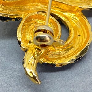 May include: Close-up of a gold-toned brooch with a swirling, textured design. The brooch features a pin closure on the back and is accented with black enamel detailing. The overall design is ornate and detailed, suggesting a vintage or antique style.
