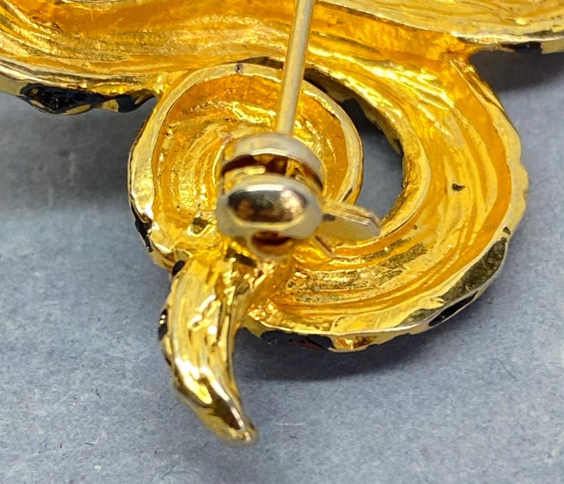 May include: Close-up of a gold-toned brooch with a swirling, textured design. The brooch features a pin closure on the back and is accented with black enamel detailing. The overall design is ornate and detailed, suggesting a vintage or antique style.