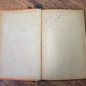 May include: Open vintage book with aged, tan pages and an orange spine. The right page has a handwritten signature and the date 11.1955. The book is open on a wooden surface.