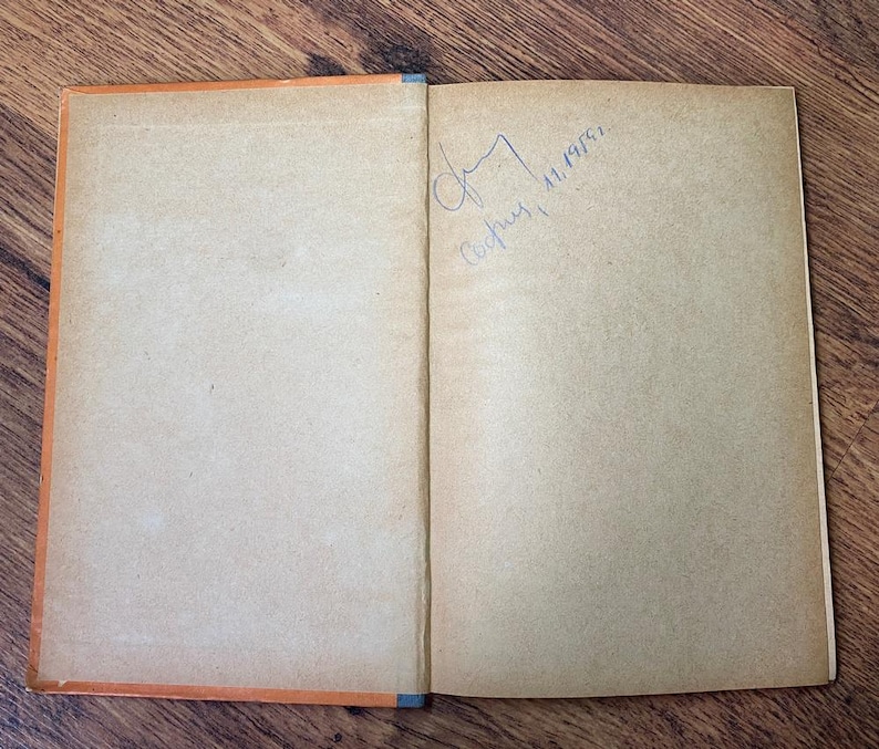 May include: Open vintage book with aged, tan pages and an orange spine. The right page has a handwritten signature and the date 11.1955. The book is open on a wooden surface.